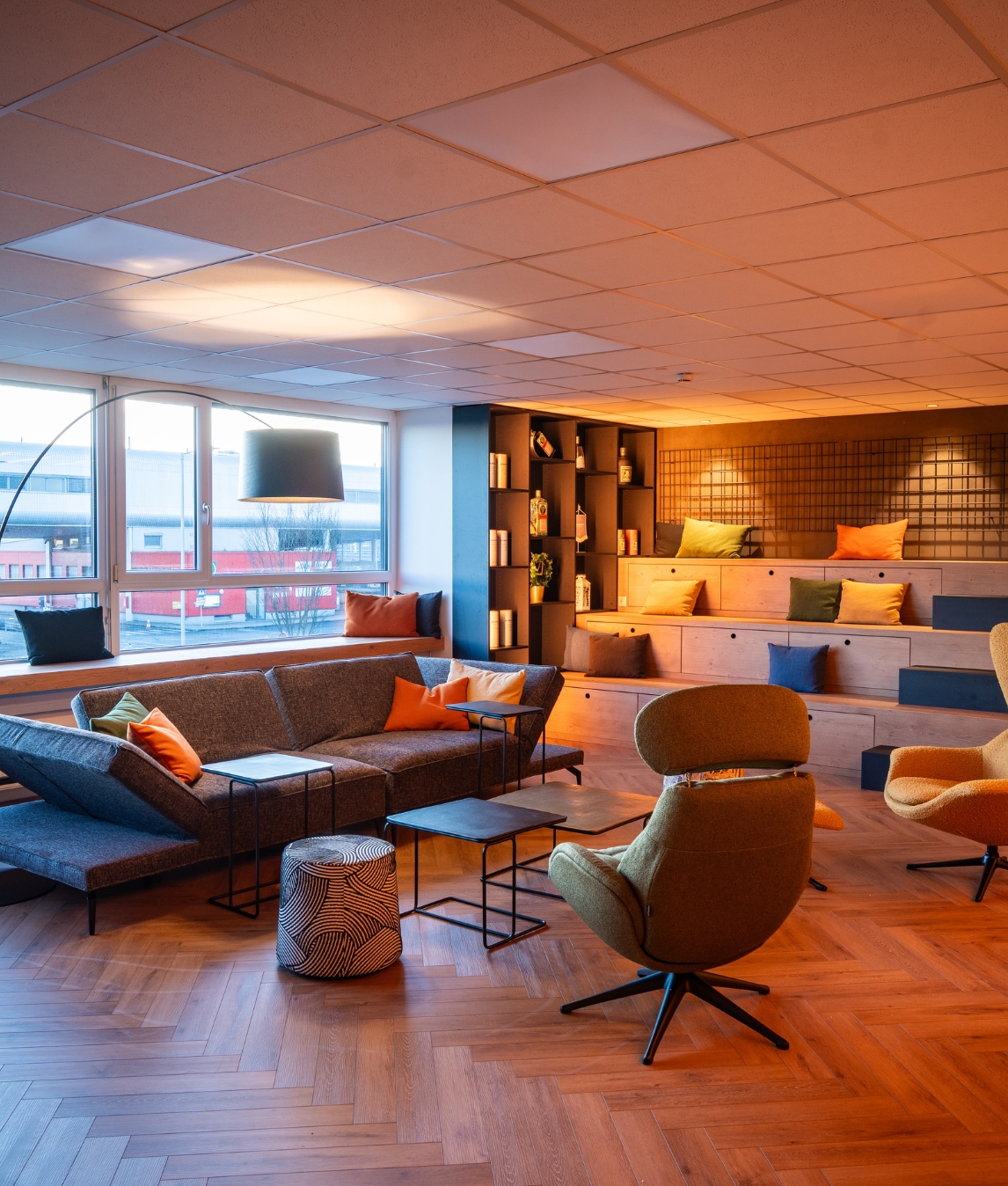 Ein modern gestalteter Aufenthaltsraum mit einem grauen Sofa, bunten Kissen in Orange und Grün, Holzboden im Fischgrätmuster und einer hölzernen Sitztreppe im Hintergrund.