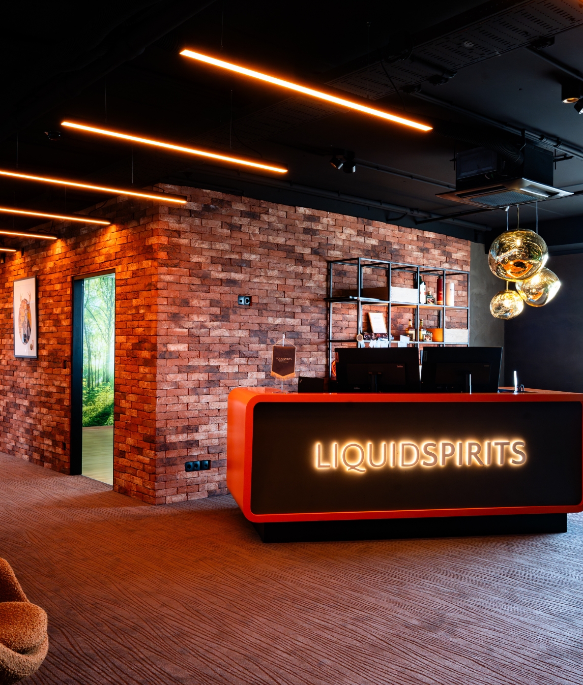 Ein moderner, orangeroter Empfangstresen mit der leuchtenden Aufschrift „LIQUIDSPIRITS“ vor einer rustikalen Backsteinwand unter industrieller Deckenbeleuchtung.
