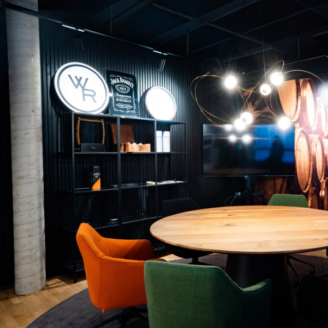 A stylish, dark room featuring a round wooden table, colorful armchairs in orange and green, and a shelf with spirits branding like Jack Daniel's.