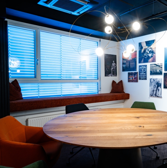 A round oak table in a bright room with large windows, blue blinds, and a cozy padded bench in the window nook.