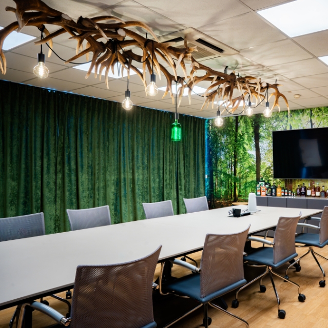 A bright conference room featuring a long white table, blue office chairs, and a forest-themed accent wall with a striking antler chandelier hanging above.
