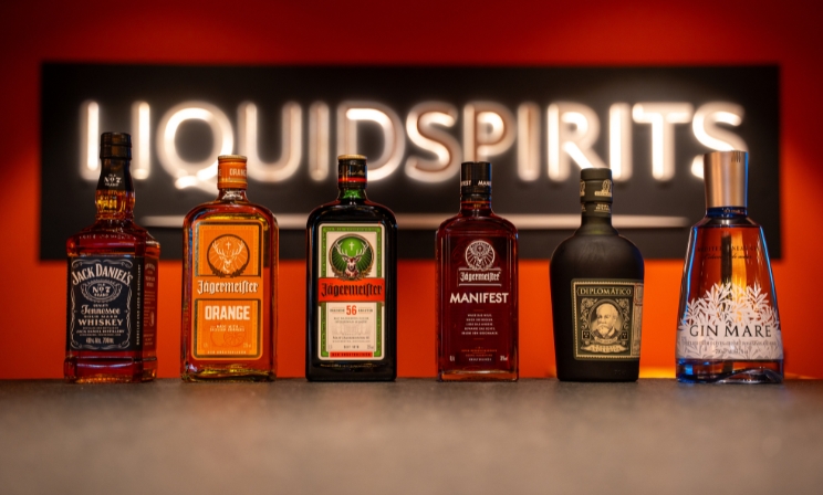 Eine Reihe verschiedener Flaschen, darunter Jack Daniel's, Jägermeister und Gin Mare, auf einem Tresen präsentiert vor dem unscharf leuchtenden LiquidSpirits-Logo.